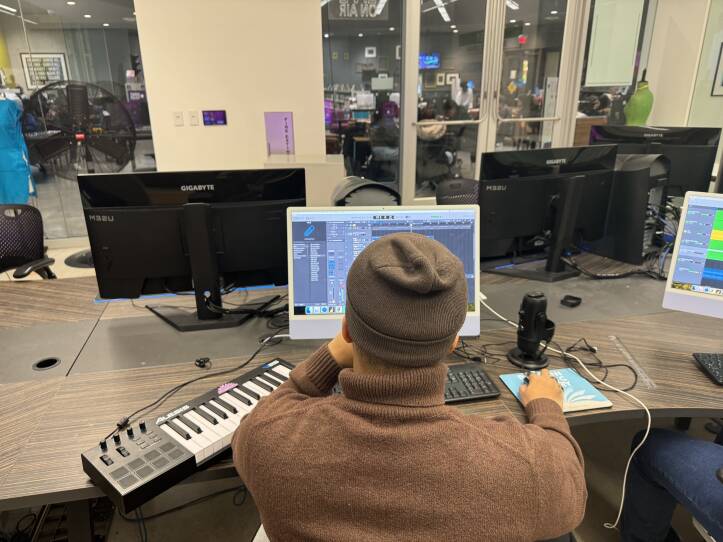 A teenage boy wearing a brown sweater and brown hat sits in front of a computer with music production software open