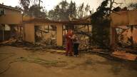 Two people embrace as they look at the rubble and char of a destroyed home.