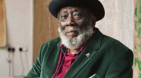 A portrait of artist Frank Bowling, wearing a green suit jacket, red button up shirt, and black hat. Bowling looks at the camera and sits in front of a beige wall.