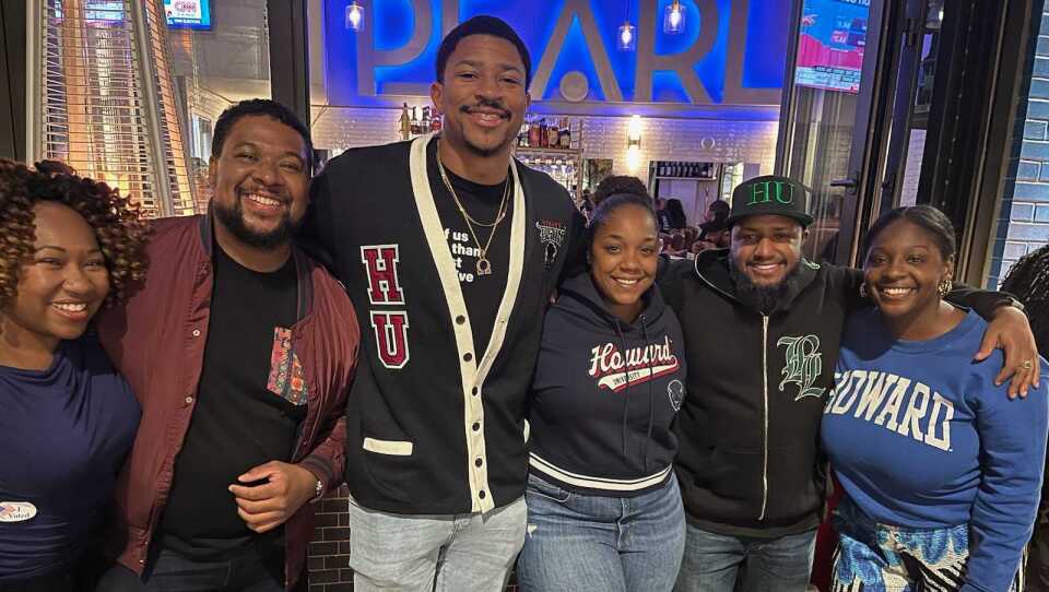 Six people stand, arms wrapped around one another, and smile at a party.