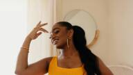 Samara Joy wears a yellow dress in a sunlit room, with her hair in a long pony tail.