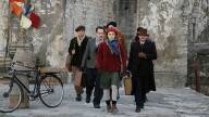 A group of men walk toward camera, led by a young woman in a red hat and coat. Behind them is a dingy stone wall.