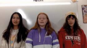 Three young women sit against a white board.