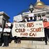 A group of people hold protest signs on the steps of the Massachusetts State House, with its gold dome visible in the background. A large banner in front says, "Leaving kids in the cold is scary! Fund EA!"