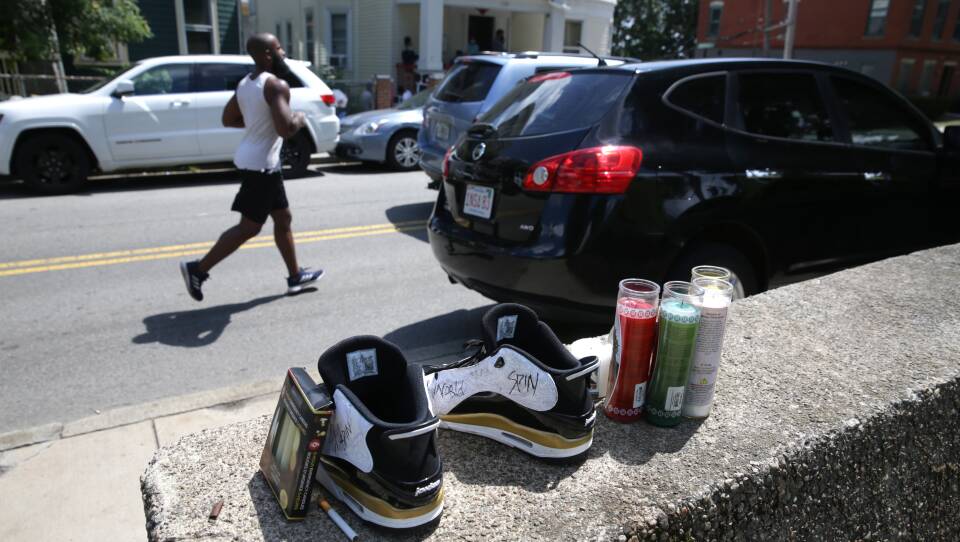 Sneakers, a pack of cigarettes and candles sit on a concrete wall. A man jobs down a street.