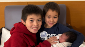 Two young boys grin while holding their baby sister in a hospital chair.