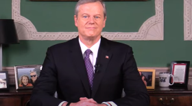Gov. Charlie Baker, in a suit and tie, sits at a desk with his hands folded and framed photos behind him.
