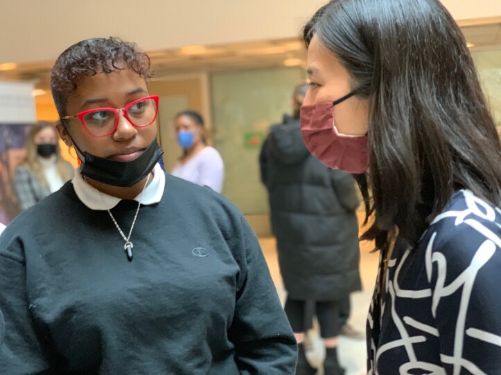 A young woman with red-framed glasses looks at another woman wearing a mask as the two speak in a classroom.