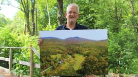 A man holds up a large photograph showing trees around a house in a valley.