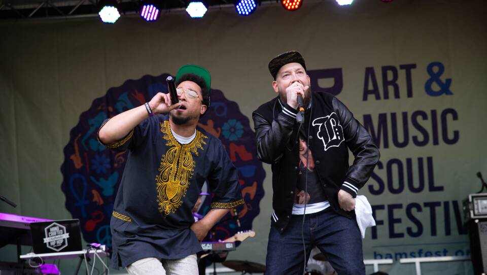 Two men with microphones on stage at BAMS Fest. One wears a black shirt with gold designs and a green hat, the other wears a black jacket that says B on it. Multicolored lights are above their heads.