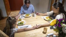 Two parents and a child sit around a dining table, holding hands as they pray before their meal.