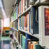 A portrait of William Shakespeare on a book cover peeks out from behind other books on ceiling-high shelves that line an aisle in a library.