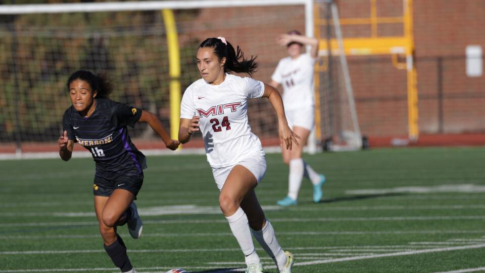 A woman wearing an MIT uniform runs down a soccer field in preparation of kicking a ball. Another player is running behind her, unable to keep up.