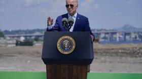 An older man in a suit stands behind a podium with the presidential seal. A barren field, river and bridge are in the background.