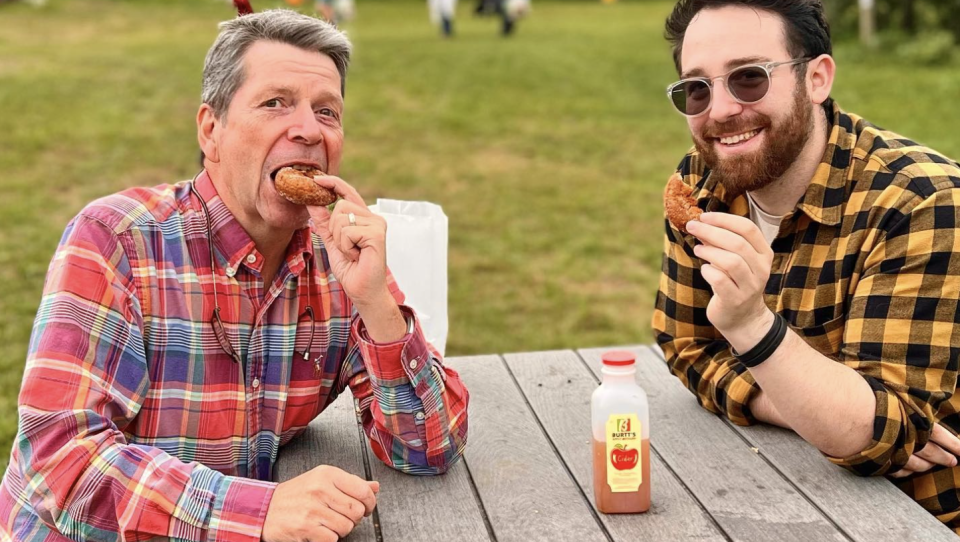 Two men wearing flannel shirts bite into apple cider donuts at an outdoor farm table