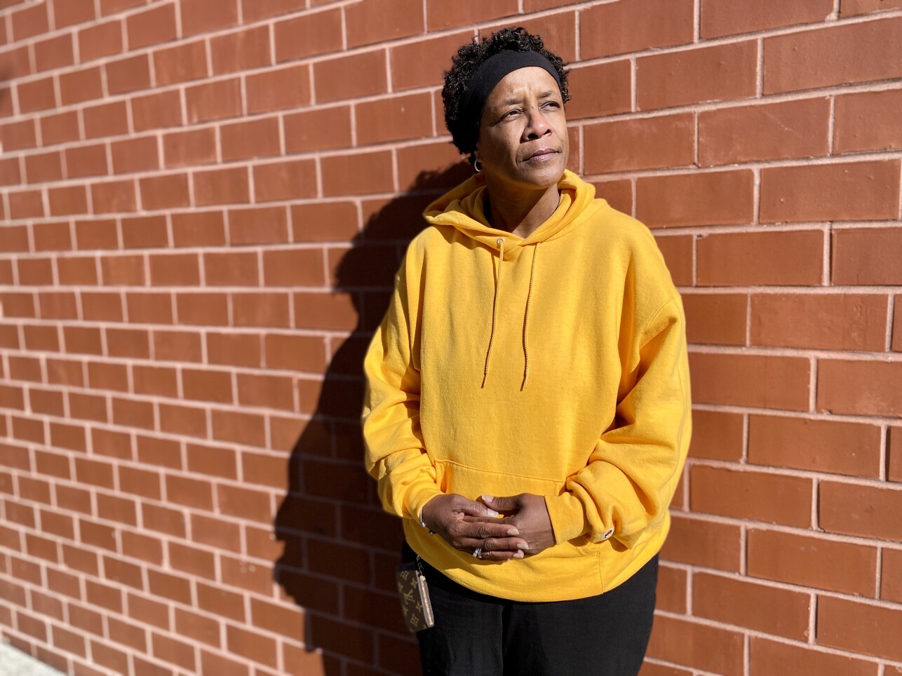 A Black woman in a yellow sweatshirt stands in front of a brick wall, the motel in Chelmsford which is her temporary shelter.