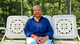A woman wearing blue pants and a blue shirt sits on a white bench surrounded by greenery.