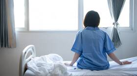 Back view of patient woman sitting on bed in hospital ward, looking away at window.