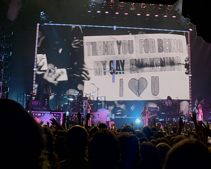 A band performs on stage. Behind them, a screen shows a sign that says 'thank you for being my gay awakening I heart you.'