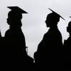 Silhouettes of four people wearing caps and gowns during an outdoor graduation ceremony.