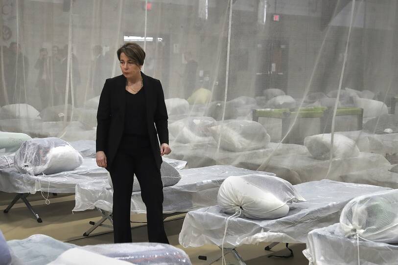 Maura Healey stands in between rows of cots covered in plastic sheeting and bags of blankets.