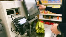 A person selects a button on the screen of a self-checkout kiosk as they weigh bananas on the scale.