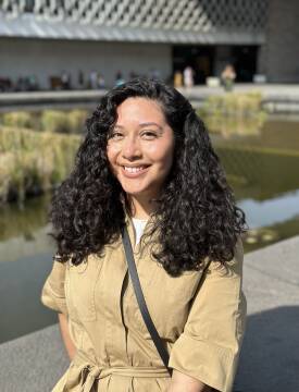 A woman sits and smiles at the camera. She wears a tan button down shirt.