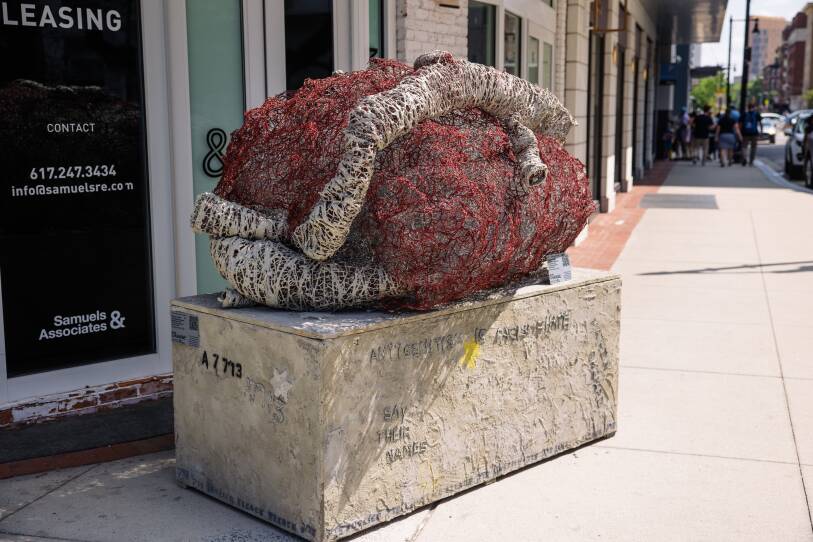This is a photo of  a public art sculpture, which is on the sidewalk. It's a rendition of a heart made from red and white chicken wire. It is mounted to a larger rectangle of concrete. It's titled  “Prisoner a-7713.”   "A-7713" is stenciled on the concrete. tihs is the  number tattooed on Holocaust survivor Elie Wiesel’s arm when he arrived in Auschwitz.