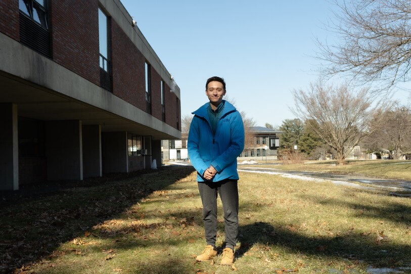 A young man in a teal jacket poses for a photo while standing on the grassy lawn outside a large building.
