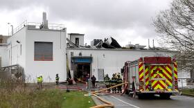 A firetruck and a half dozen firefighters are outside a factory with rubble on top.