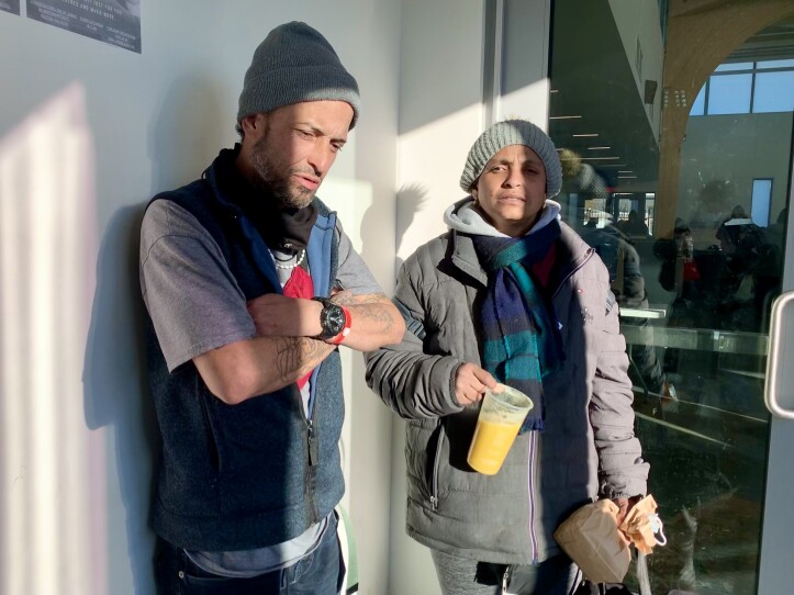 The couple stands in a vestibule, both wearing winter hats. The man looks down with his arms crossed, mid-speech.