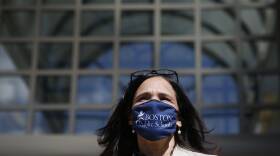 A woman wearings a "Boston Public Schools" mask and glasses atop her head stands outside a large building.