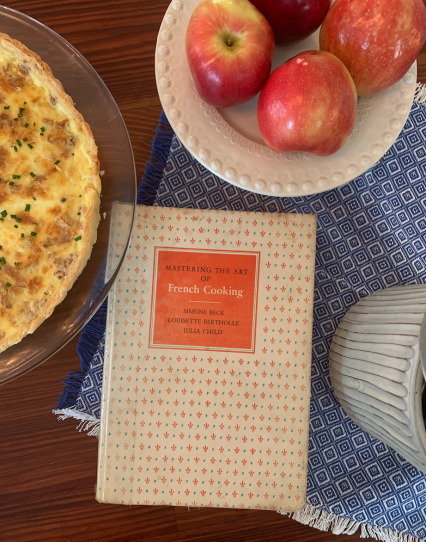 An cookbook sits on a kitchen table, surrounded by a bowl of apples and a quiche
