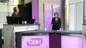 A woman in a headset stands at a purple-lit desk. To her left is a man in a headset, behind a computer.