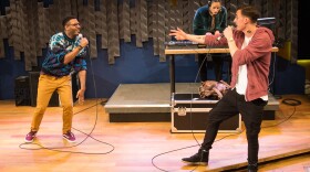 Three people are on a stage performing a hip-hop song. Two men face each other, holding microphones as they sing. in the middle is a young woman DJ, bending over a mixing console