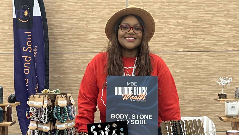 A woman wearing a hat and a beaded bracelet smiles as she stands behind a table containing plenty of bracelets, rings, necklaces and other goods.