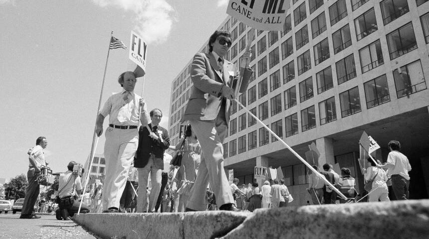 Several men using canes walk on a raised sidewalk outside a large government building.