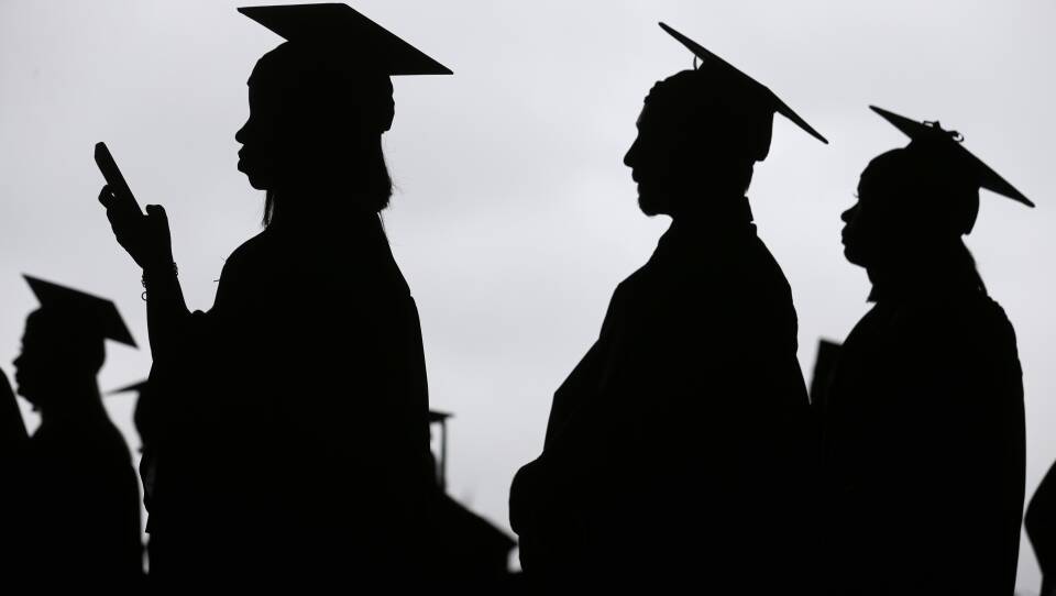 Silhouettes of four people wearing caps and gowns during an outdoor graduation ceremony.