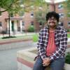 A young Hispanic man sits smiling outside of his public housing complex in Somerville. He has curly dark brown hair, cropped in on the sides, that piles up on the top of his head, and he smiles at the camera underneath his mustache and black plastic glasses, his hands clasped in his lap.