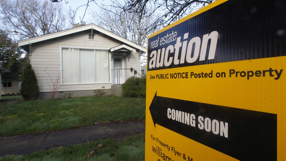 A yellow yard sign is posted on the grass outside a small house. The sign says "real estate auction coming soon. See public notice posted on property."