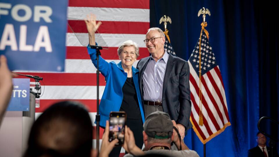 Elizabeth Warren smiles and waves while standing on stage in front of an American Flag backdrop. Her partner has his arm wrapped around her.