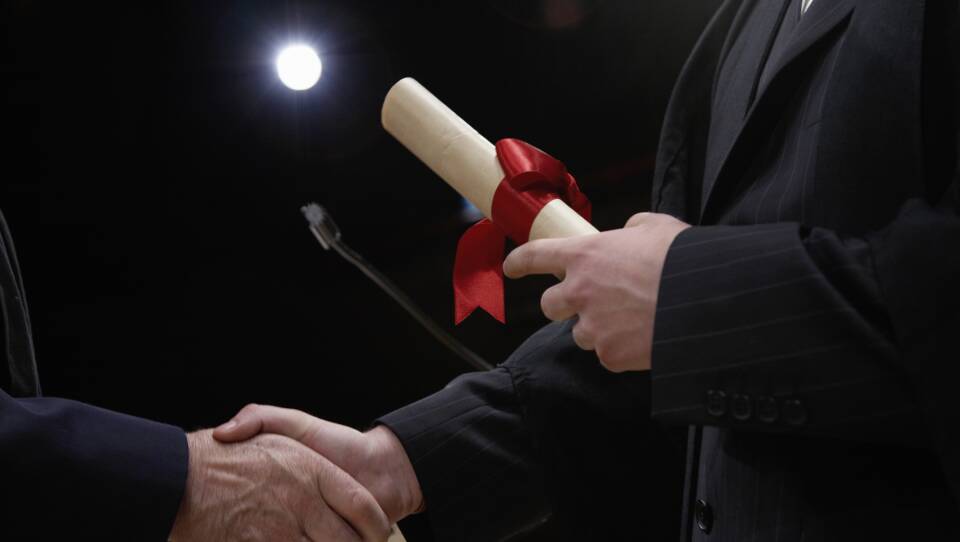 A person shakes another person's hand as they hold onto a rolled-up sheet of paper with a ribbon tied around it.