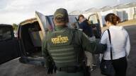 A person wearing a CBP Border Patrol Federal Agent uniform holds a woman by the arm as they lead her to a van where a couple other people are.