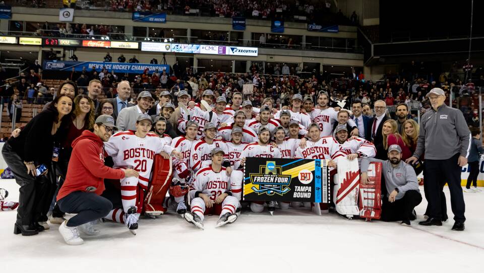 A men's ice hockey team in full red-and-white uniforms pose on an ice rink with a banner reading "frozen four Tampa bound."
