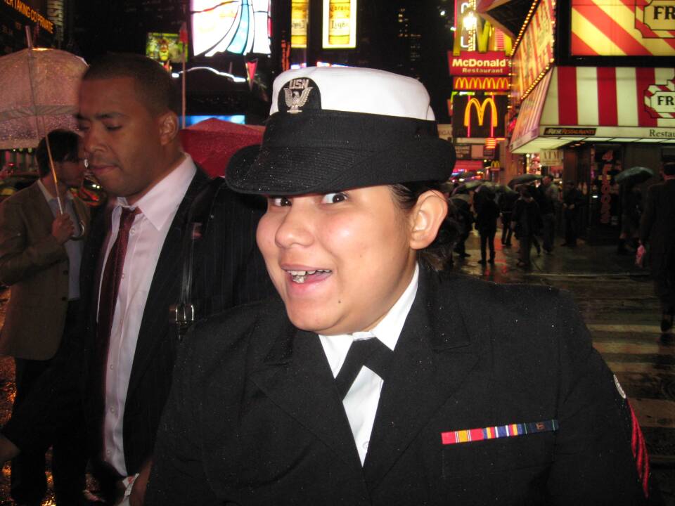 A smiling woman wearing a U.S. Navy cap and uniform stands on the sidewalk of a city street at night.