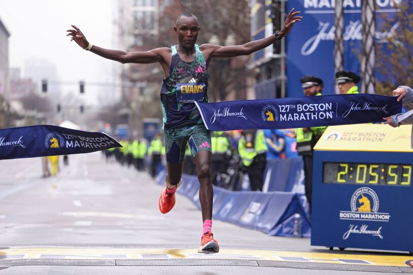 Chebet runs through the tape at the Boston Marathon finish line, his hands out to his side. It's a gray cloudy day.