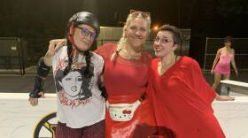 Three women pose for a photo. One is wearing leopard print shorts and a punk t-shirt, the other two are in all red.