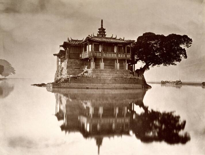 This is a photograph taken around 1871 of a pagoda in he city of Fuzhou (Foochow) in Fujian province on the River Min. The photo is in sepia tone. A pagoda appears to be floating on a still river. It's reflected in the river. There are a couple of fishing boats in the background.
