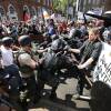 Dozens of protesters and counter-protesters clash in the street. Some hold signs, other wave flags, others still appear to be wearing helmets or body armor. Brick buildings on a city street stand in the backdrop.