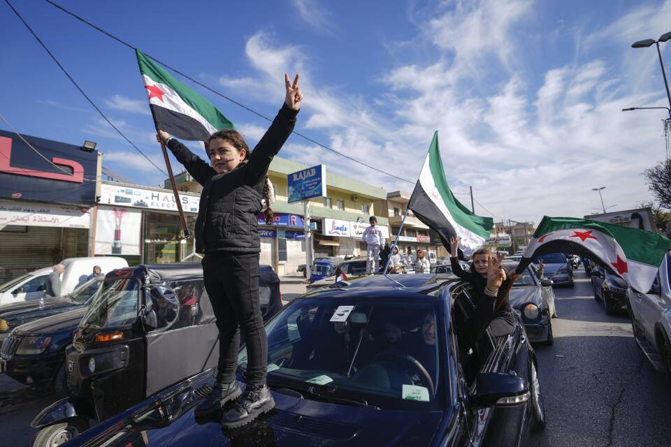 A kid waving a Syrian flag stands on the hood of a car
